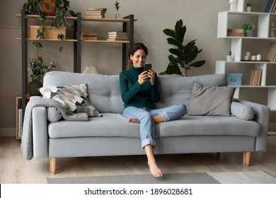 Smiling young Caucasian woman sit relax on couch in living room talk on video call. Happy millennial female rest on sofa at home have webcam digital virtual communication on cellphone.