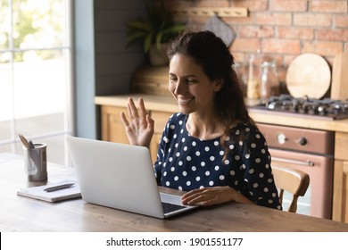 Smiling young Caucasian woman sit at table at home kitchen wave greet talking on video call on computer gadget. Happy millennial female look at laptop have webcam online digital lesson or training.