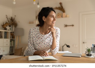 Smiling young Caucasian woman sit at table at home write make list in notebook look in distance dreaming thinking. Happy millennial female handwrite in notepad visualize or imagine. Planning concept.