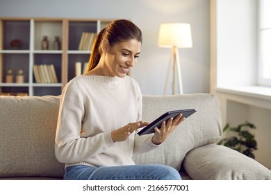 Smiling Young Caucasian Woman Sit On Sofa Use Modern Tablet Gadget. Happy Millennial Girl Relax At Home Shopping Online Or Browse Internet On Pad Device. Technology And Communication.