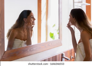 Smiling Young Caucasian Woman In Towel Look In Mirror In Home Bath Apply Facial Moisturizing Cream Or Serum, Happy Millennial Female Perform Daily Face Skincare Routine Or Treatment In Bathroom