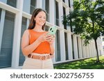 Smiling young Caucasian woman using her smartphone with a happy face standing outside at the university campus. Pretty teenage college student reading text messages, browsing or chatting on cellphone