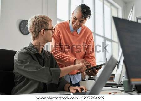 Smiling young colleague consulting with programmer about new software on digital tablet during their work in office
