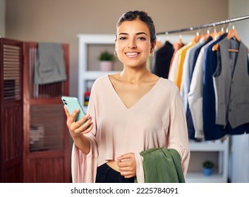 Smiling Young Confident Indian Asian female professional fashion Designer holding a Mobile phone and dress outfit in hand in boutique looking at camera. Self-employment, New Business Startup concept.