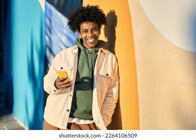 Smiling young cool African American guy holding mobile phone tech device standing at colorful city wall. Happy stylish authentic hipster teen boy using apps on cell outdoors, looking at camera.