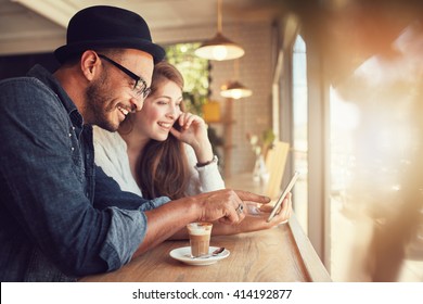 Smiling Young Couple In A Coffee Shop Using Touch Screen Computer. Young Man And Woman In A Restaurant Looking At Digital Tablet.