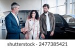Smiling young couple discussing automobile specifications with salesman, while visiting car dealership. Happy husband and wife using consultation of auto dealer with tablet while choosing car