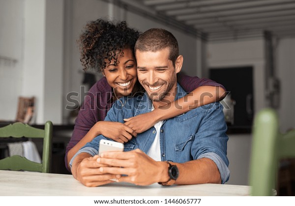 スマートフォンを見ながら抱きしめながら微笑む若い夫婦 スマートフォンでソーシャルメディアを共有する多民族カップル 携帯電話を使いながら 幸せな彼氏の後ろから抱きしめる笑顔のアフリカの女の子 の写真素材 今すぐ編集