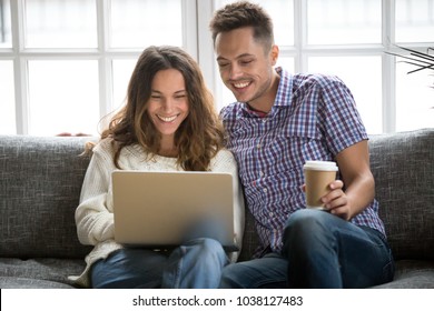 Smiling Young Couple Enjoying Morning Coffee With Computer Sitting On Sofa At Home Together, Happy Man And Woman Laughing Looking On Laptop Screen Having Fun Or Calling Online, Watching Funny Video