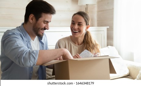 Smiling young couple feel overjoyed unpack cardboard package ordering buying goods over internet use delivery service, happy millennial husband and wife customers open box purchase shopping online