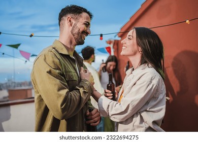 Smiling young couple flirting and having fun at a rooftop party with friends, drinking beer, enjoying and having a pleasant conversation. Handsome guy greeting his joyful girlfriend at celebration - Powered by Shutterstock