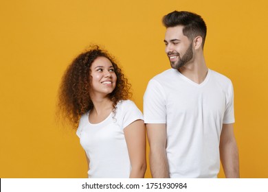 Smiling Young Couple Friends European Guy African American Girl In White T-shirts Isolated On Yellow Background. People Lifestyle Concept. Mock Up Copy Space. Stand Back To Back Looking At Each Other