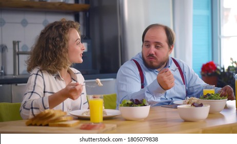 Smiling Young Couple Having Breakfast In Kitchen At Home. Overweight Guy And Attractive Woman Eating Tasty Dinner Together At Home. Healthy Diet, Weight Loss Concept