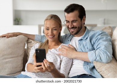 Smiling young couple hugging using smartphone sitting on cozy couch together, happy woman and man in glasses looking at phone screen, watching video, chatting, satisfied clients shopping online