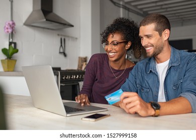 Smiling Young Couple Making Shopping Online With Credit Card And Laptop At Home. Happy Multiethnic Couple Holding Debit Card While Buying On Ecommerce Site Using Laptop. 