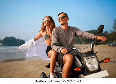 Smiling young couple ride motorbike along tropical beach. Woman spreads arms in freedom, man steers scooter. Adventure vacation ride, enjoy sea breeze. Happy pair exploring travel destination.