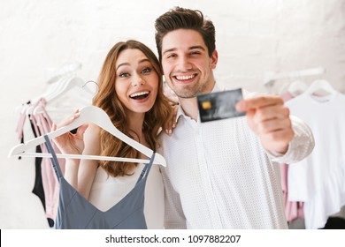 Smiling Young Couple Shopping For Clothes Together At The Clothing Store, Woman Holding Dress On A Hanger And Man Showing Credit Card