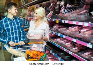 Smiling young couple with trolley choosing meat in supermarket