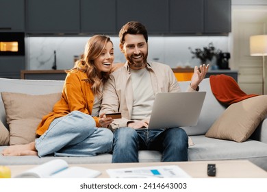 Smiling young couple using laptop computer holding credit card shopping online sitting on sofa at home. Attractive man and woman, freelancer working online receive payment. Successful business  - Powered by Shutterstock