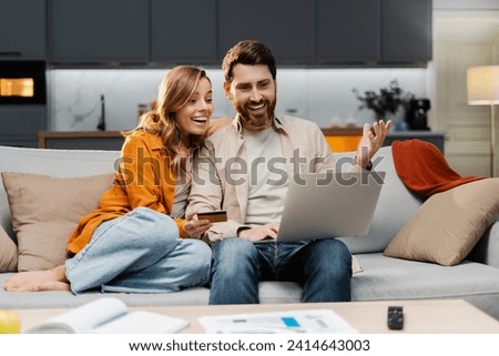 Smiling young couple using laptop computer holding credit card shopping online sitting on sofa at home. Attractive man and woman, freelancer working online receive payment. Successful business 