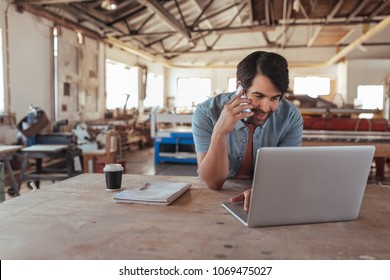 Smiling young craftsman standing at a workbench in his large workshop working online with a laptop and discussing designs with a customer on a cellphone - Powered by Shutterstock