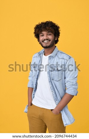 Smiling young curly indian cool positive guy standing isolated on yellow background. Happy ethnic stylish millennial man wearing blue casual t-shirt looking at camera posing for portrait. Vertical