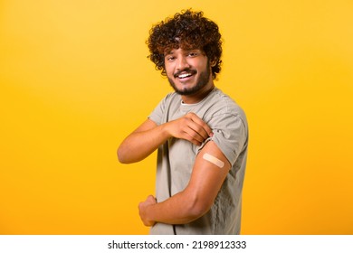 Smiling Young Curly Indian Guy Showing Arm With Band-aid After Vaccine Injection Isolated On Yellow Background, Vaccinated Multiracial Man Feels Safe With Vaccine