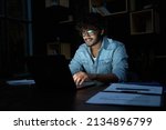 Smiling young curly indian latin ethnic business man or student wearing glasses remote working overtime, learning online late at night at home or in dark office using laptop computer at workplace.