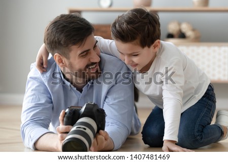 Smiling young dad lying on floor with preschooler son holding professional camera show pictures to child, happy father photographer and boy kid making pictures together with photographic device