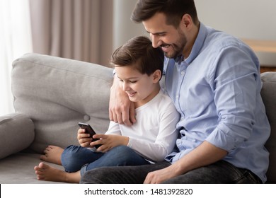 Smiling Young Dad Sitting On Couch With Pretty Preschool Son Watching Funny Videos On Smart Phone Using New App, Happy Father And Kid Relaxing At Home Play Games On Cellphone Modern Tech Usage Concept