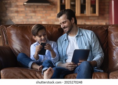 Smiling Young Dad And Small Son Relax On Sofa At Home Use Modern Electronic Devices Together. Happy Caucasian Father Rest On Couch In Living Room With Teen Boy Child Browsing Internet On Gadgets.