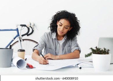 Smiling young dark-skinned self-employed woman architect with Afro hairstyle making drawing at her home office using pen, excited about working process. Engineering, construction and architecture - Powered by Shutterstock