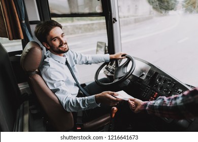 Smiling Young Driver taking Ticket from Passenger. Handsome Happy Man wearing Blue Shirt Sitting on Driver Seat of Tour Bus. Attractive Confident Man at Work. Traveling, Transport and Tourism Concept