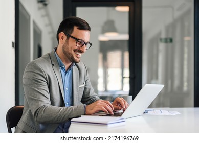 Smiling young entrepreneur typing on laptop. Happy businessman is working over strategy at desk. He is wearing eyeglasses while planning at office. - Powered by Shutterstock