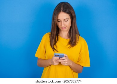 Smiling Young european brunette woman wearing yellow T-shirt on blue backgroundusing cell phone, messaging, being happy to text with friends, looking at smartphone. Modern technologies and communicati