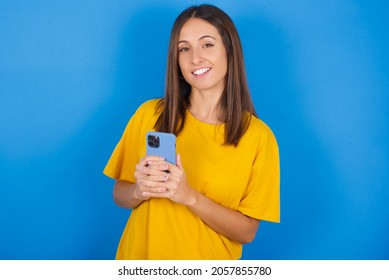 Smiling Young European Brunette Woman Wearing Yellow T-shirt On Blue Background Friendly And Happily Holding Mobile Phone Taking Selfie In Mirror.