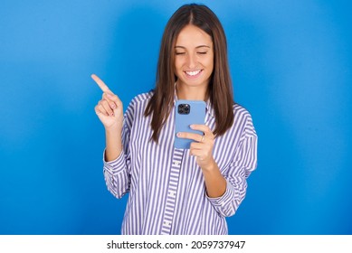 Smiling Young European Brunette Woman Wearing Stripped Shirt On Blue Background Pointing Finger At Blank Space Holding Phone In One Hand