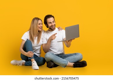 Smiling young european guy with beard and lady sit on floor, show finger in laptop isolated on yellow background, studio. Relationships, video call, meeting remotely and new normal, tech at covid-19