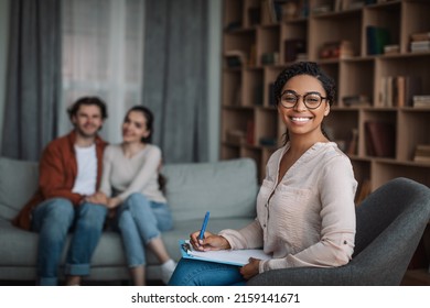 Smiling young european guy and lady sit on sofa at meeting african american woman doctor psychologist in office interior. Consultation, professional help, support, family psychology, relationships - Shutterstock ID 2159141671