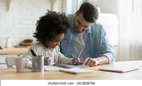 Smiling young european man helping little adopted african american daughter with homework, sitting together at table. Happy small biracial child girl involved in doing school tasks with foster father.