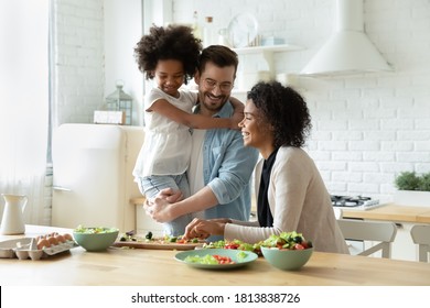 Smiling young european man holding on hands pretty small biracial adopted girl daughter, watching happy african american mother preparing healthy food, chopping fresh vegetables for salad in kitchen. - Powered by Shutterstock