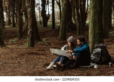 Smiling young european man and woman tourists in jackets with backpack resting in autumn forest, enjoy cold season outdoors, looking at map with hot drink from thermos. Route for travel and nature - Powered by Shutterstock