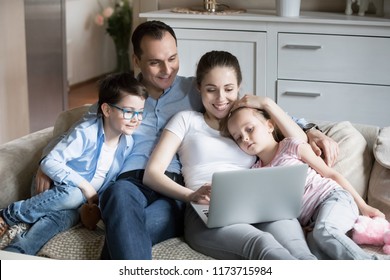Smiling young family lying with kids on cozy sofa watching cartoons on laptop, happy parents hug son and daughter relaxing on couch with computer, mom and dad spend time with children at home