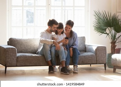 Smiling young family with preschooler child have fun using smartphone relaxing on couch, happy little girl hold cellphone play game enjoy spending time at home, rest on sofa with mom and dad
