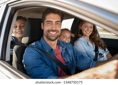 Smiling Young Family With Two Children Sitting In Car And Driving. Family Relaxing During Road Trip While Looking At Camera. Portrait Of Happy Father Riding In A Car With Wife, Son And Daughter.