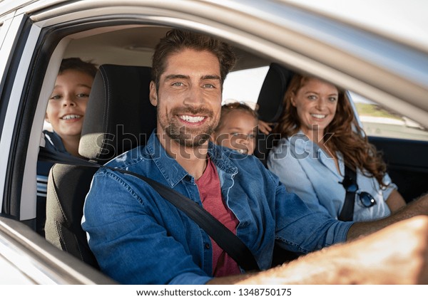 Smiling young family with two children sitting\
in car and driving. Family relaxing during road trip while looking\
at camera. Portrait of happy father riding in a car with wife, son\
and daughter.