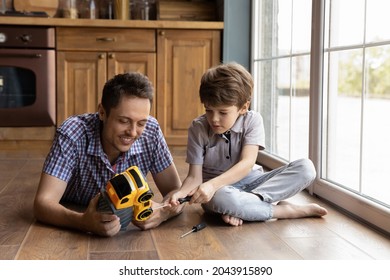 Smiling young father teaching little son repairing toy, playing together on warm floor in kitchen. Adorable small concentrated kid boy fixing car using screwdriver, improving skills with dad at home.