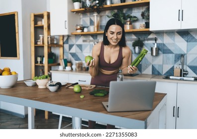 Smiling young female in activewear with celery and apple cooking healthy food while watching online cooking class on laptop in contemporary kitchen