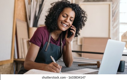 Smiling young female artisan working on a laptop and talking with a client on the phone in her workshop - Powered by Shutterstock