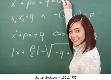 Smiling young female Asian student or teacher doing maths standing at a blackboard covered in mathematical equations